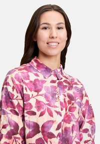 Koszula w kwiaty z fioletowymi i różowymi wzorami na kremowym tle. Materiał jest teksturowany, z zapięciem na guziki i kołnierzykiem.