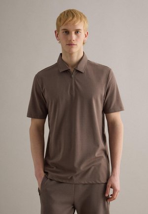 Junger Mann mit kurzen blonden Haaren, der ein braunes kurzärmeliges Polohemd mit Reißverschluss und passende Hose trägt, vor einem einfarbigen Hintergrund stehend.