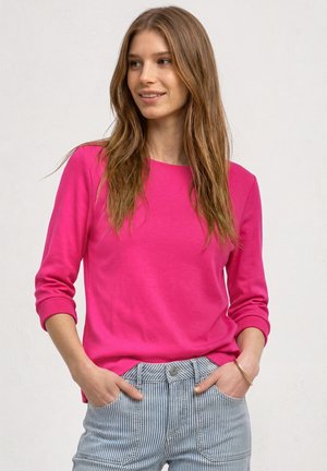 Jonge vrouw met lang bruin haar, die een felroze shirt met lange mouwen en lichtblauwe gestreepte broek draagt, handen in de zakken, licht glimlachend.