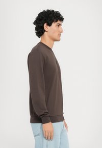 Felpa marrone con maniche lunghe, scollatura rotonda, polsini a costine e orlo aderente. Abbinata a jeans di denim azzurri chiari.