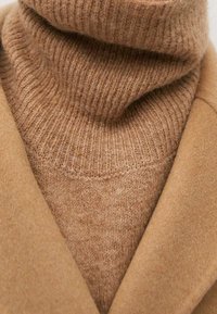 Pull à col roulé couleur fauve en maille côtelée, superposé sous un manteau beige sur mesure. Texture douce avec une finition lisse et des lignes épurées.