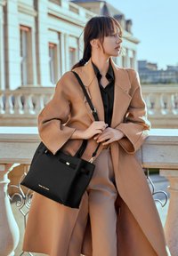 Zwart leren handtas met een gestructureerde vorm, gouden accenten en dubbele handvatten. Gecombineerd met een beige wollen jas en lichtbruine broek.