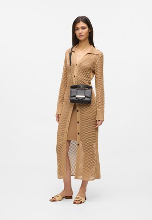 Vrouw met een lange beige mesh cardigan over een bijpassende jurk, met een zwarte crossbodytas en beige open-toe sandalen met lage hak.