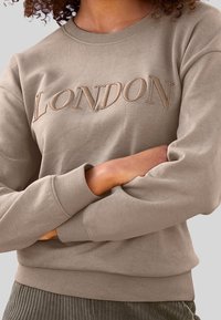 Béžová mikina s výšivkou "LONDON" v tlumených tónech, má žebrované manžety a spodní lem a klasický kulatý výstřih.