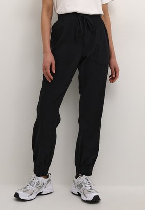 Personne portant un pantalon jogger noir avec des poignets élastiques, des baskets blanches et un t-shirt blanc rentré, debout contre un mur uni.
