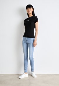 Camiseta de algodón negra de manga corta con el logo "Tommy", combinada con jeans ajustados de color azul claro y zapatillas blancas, de pie contra un fondo neutral.
