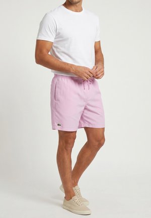 Hombre con camiseta blanca, pantalones cortos rosa claro con cordón y logo, y zapatillas beige con cordones, de pie sobre un fondo claro y liso.