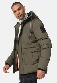 Doudoune verte olive avec capuche, comportant une fermeture éclair à l'avant, plusieurs poches, un patch texturé sur la manche et des accents avec cordon de serrage.