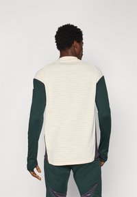 Long-sleeve athletic shirt with a cream body, dark green sleeves, and a purple accent. Textured fabric with a subtle pattern.