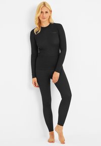 Blonde woman wearing a fitted black long-sleeve top and leggings, standing barefoot against a white wall, relaxed pose.