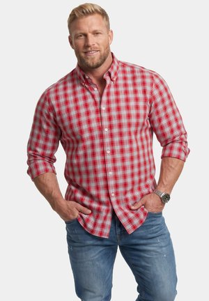 Blonder Mann mit Bart trägt ein rot-weiß kariertes Hemd und blaue Jeans, Hände in den Taschen, lächelt in die Kamera vor weißem Hintergrund.