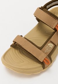 Rjave in peščene sandalske čevlje z nastavljivimi tekstilnimi trakovi, teksturirano gumijasto podplato in oranžnim dodatkom. Značka se ponaša z logotipom North Face.