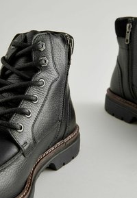 Botas de cuero negro con acabado texturizado, que presentan ojales de metal, cordones, cremalleras laterales y una suela robusta con un detalle de costura en contraste.