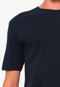 Nærbilde av en person som har på seg en marineblå ribbestrikket kortermet skjorte med rund hals mot en ensfarget hvit bakgrunn.