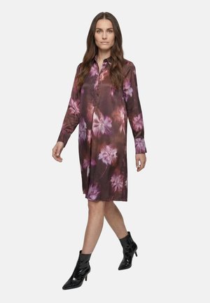 Frau geht und trägt ein langärmeliges, knielanges lila-braunes Blumenkleid mit schwarzen Stiefeletten vor weißem Hintergrund.