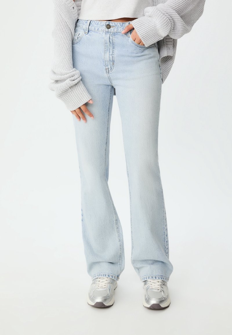 Personne portant un jean évasé bleu clair, des baskets argentées, un top court blanc et un cardigan tricoté gris oversize avec les mains dans les poches.