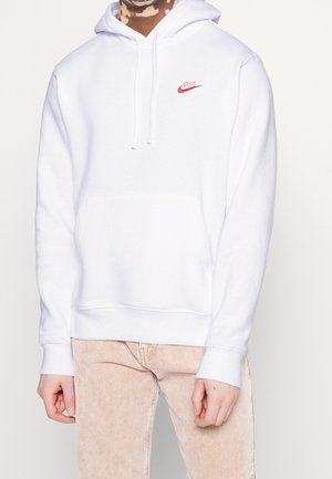 Persoon draagt een witte Nike-hoodie met rood logo op de borst en lichtbeige corduroy broek, staand tegen een effen witte achtergrond.