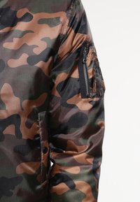 Kamuflāžas jaka zaļā, brūnā un melnā krāsā. Ar rāvējslēdzēja kabatu kreisajā piedurknē, izgatavota no gludas, spīdīgas auduma.