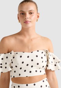 Nuori nainen, joka on pukeutunut valkoiseen off-shoulder crop topiin, jossa on mustia pilkkuja, ja kultaisiin renkaisiin, yksinkertaista taustaa vasten.