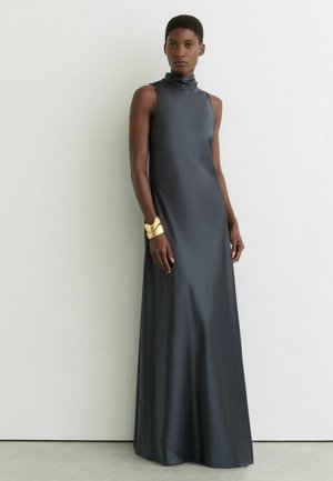 Naine seisab valge seina taustal, kandes varrukateta tumehalli maani ulatuvat kleiti ja suurt kuldset käerauakujulist käevõru vasakul randmel.