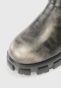 Zbliżenie na palce czarnego skórzanego buta z teksturowaną, grubą gumową podeszwą i widocznym szwem na jasnym tle.