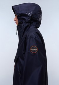 Chaqueta impermeable en color marino con capucha completa, bolsillos con cremallera y un parche con el logotipo en la manga; tejido sintético suave con un diseño elegante.