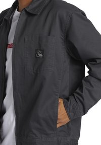 Gray jacket made of fabric with a smooth texture, featuring a front pocket and a small logo patch on the chest.