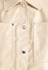 Beżowa koszula z eko skóry z klasycznym kołnierzykiem, wyposażona w srebrne zatrzaski oraz dwie kieszenie na piersi, o gładkim, teksturowanym wykończeniu.