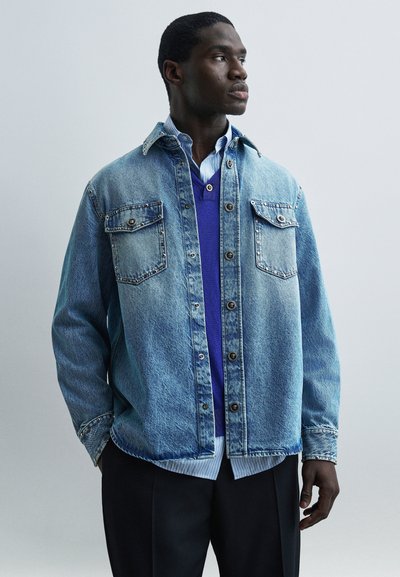 Homme portant une veste en jean bleue par-dessus un pull bleu et une chemise rayée, regardant vers la droite avec les mains dans les poches, sur un fond uni.