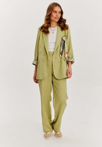 Jasnozielona, dwurzędowa marynarka z podwiniętymi rękawami, w komplecie z szerokimi spodniami i białą bluzką. Dodatki to wzorzysty szalik.