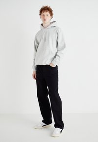 Haut à capuche gris avec poche kangourou, pantalon noir et baskets blanches avec accents bleu marine. Le mannequin se tient sur un fond neutre, les mains dans les poches.