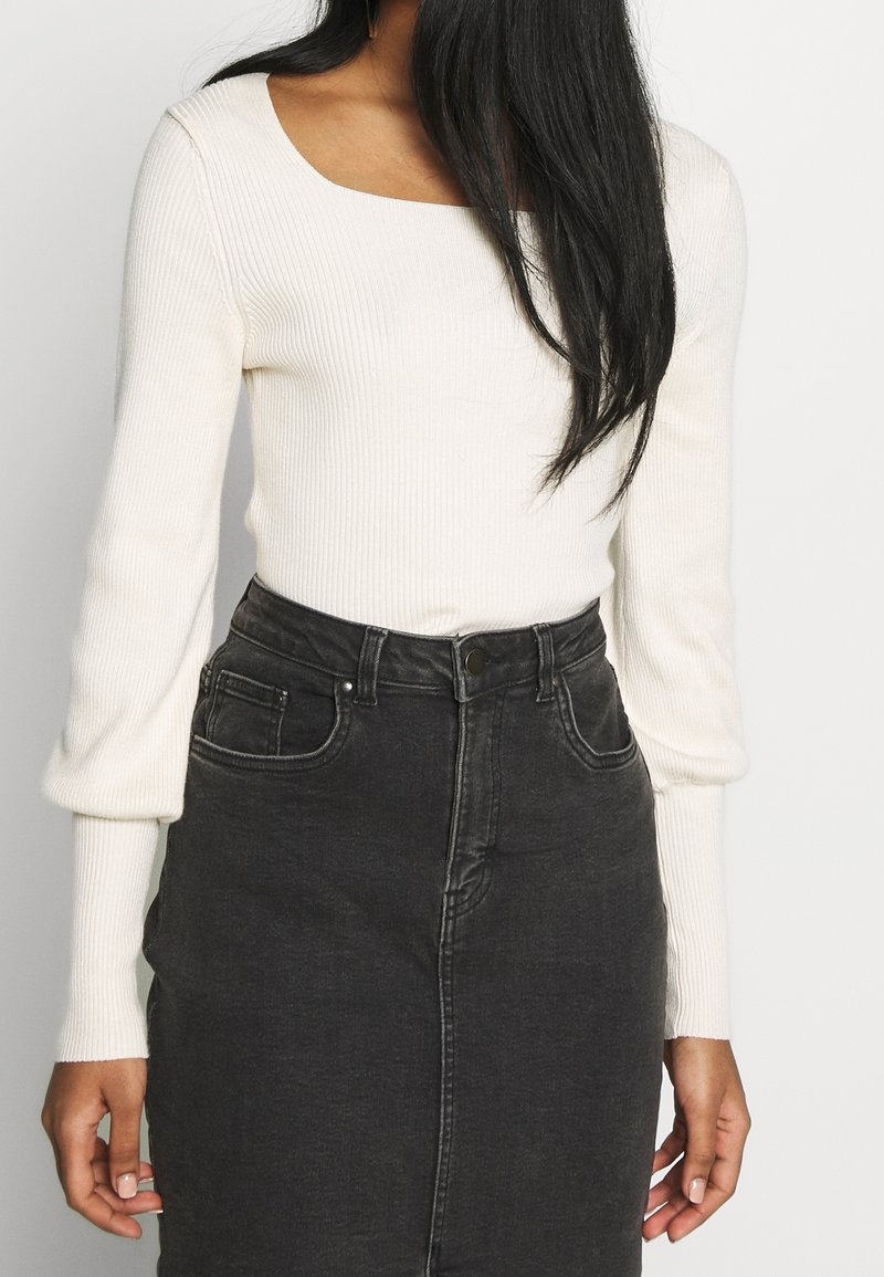 Femme portant un haut blanc moulant en maille côtelée à manches longues bouffantes, rentré dans une jupe en jean noire taille haute.