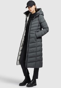 Manteau d'hiver gris avec capuche, présentant un design matelassé, une fermeture éclair à l'avant, des poches latérales et une doublure intérieure légère. Porté avec un pantalon noir et des bottes.