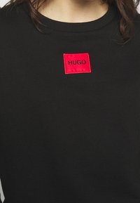 Černá mikina vyrobená z bavlny s kulatým výstřihem, na hrudi se nachází jasně červený obdélníkový nášivka se slovy "HUGO" v černém písmu.