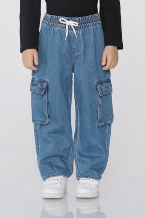 Enfant portant un pantalon cargo ample en denim bleu avec une taille élastique et un cordon blanc, associé à des baskets blanches et un haut noir à manches longues.