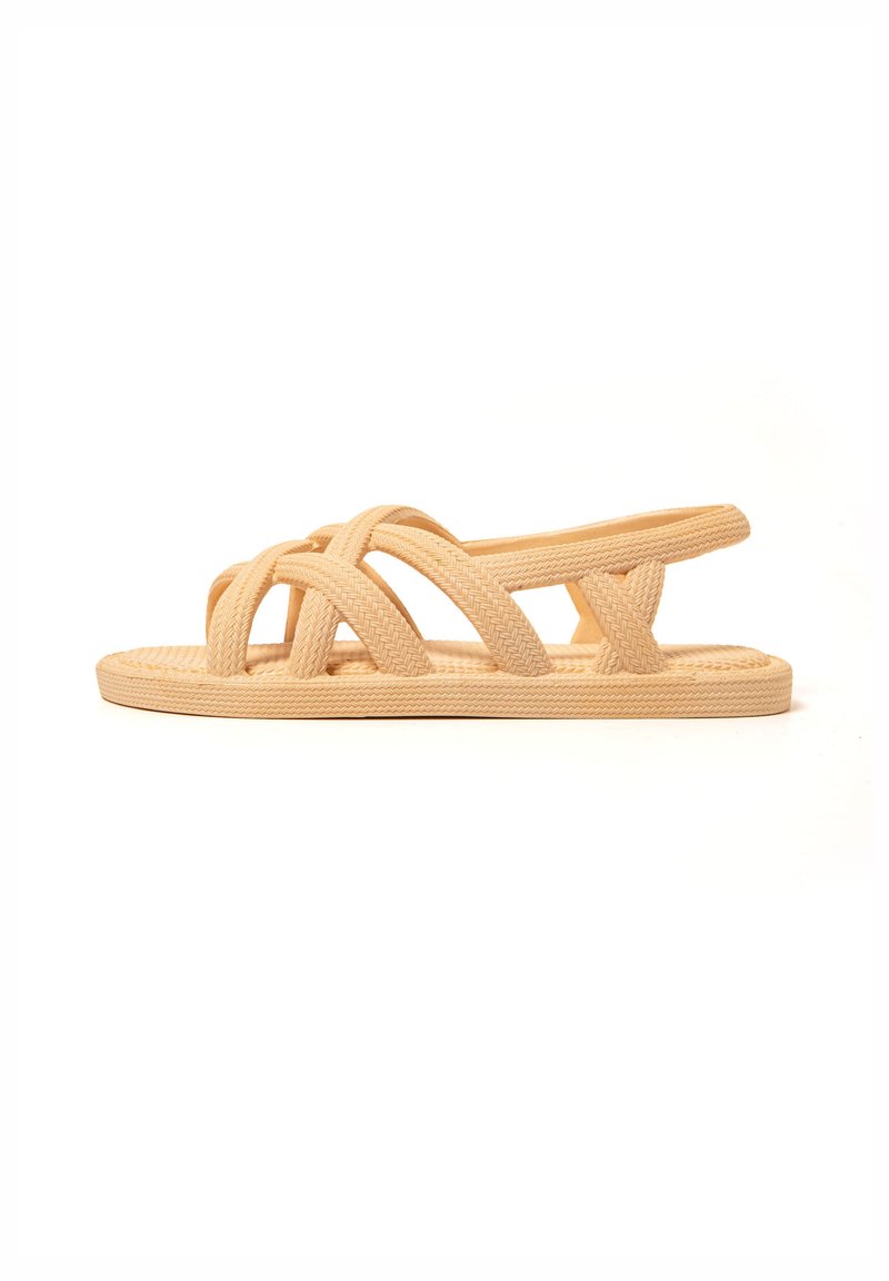 Tan woven slide sandal featuring crisscross straps, flat sole, and open heel design. Made from flexible material for comfort and breathability.