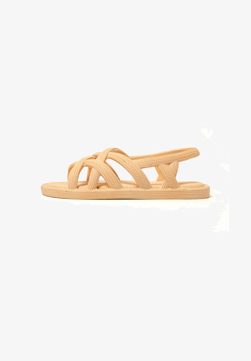 Tan woven slide sandal featuring crisscross straps, flat sole, and open heel design. Made from flexible material for comfort and breathability.