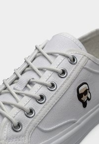 Λευκό πάνινο αθλητικό παπούτσι με λευκά κορδόνια περασμένα από μεταλλικά πόρπες, με ένα μικρό λογότυπο ενός ατόμου με γυαλιά ηλίου στο πλάι.