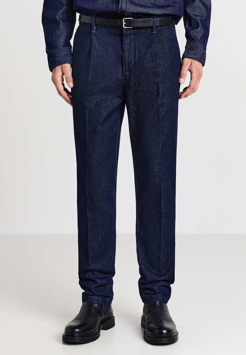 Pantaloni in denim blu scuro con vestibilità dritta, caratterizzati da pieghe frontali, tasche laterali e una cintura nera. Texture liscia e linee pulite.