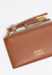 Rudas odinis pinigine su tekstūruota paviršiumi, auksine užtrauktuku ir pintu dirželiu, dekoruota iškilmingu prekės ženklo logotipu "BIMBA Y LOLA."
