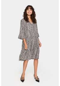 Musterkleid mit V-Ausschnitt und dreiviertel langen Glockenärmeln. Verfügt über ein schwarz-beiges Zickzack-Muster. Getragen mit schwarzen Riemchenschuhen.