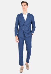 Abito blu a doppio petto con revers a scollo a V, tessuto testurizzato, pantaloni slim fit e mocassini beige. Camicia bianca sotto la giacca dell'abito.