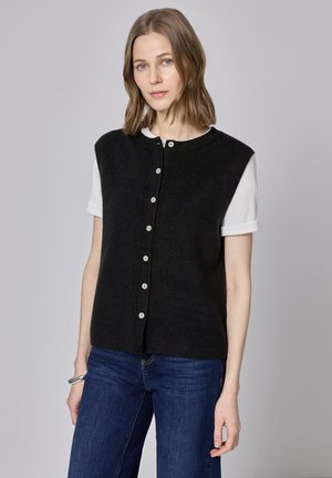 Gilet nero lavorato a maglia con maniche corte, caratterizzato da una chiusura frontale con bottoni e una texture a coste. Abbinato a una camicia bianca sotto e jeans blu.