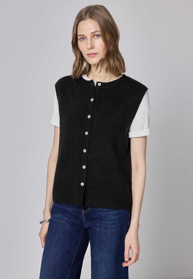 Schwarze Strickweste mit kurzen Ärmeln, vorne mit Knöpfen und einer gerippten Struktur. Kombiniert mit einem weißen Hemd darunter und blauen Jeans.