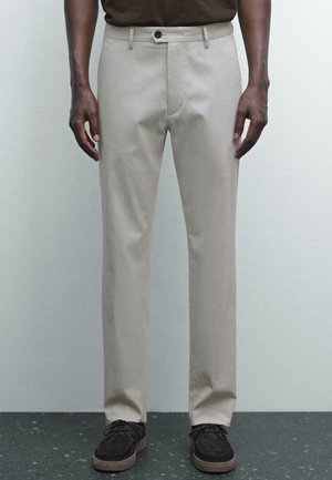 Beige Hosen mit schlankem Schnitt, flacher Vorderseite und Einzelknopfverschluss. Getragen mit dunklem Schuhwerk, vor neutralem Hintergrund.