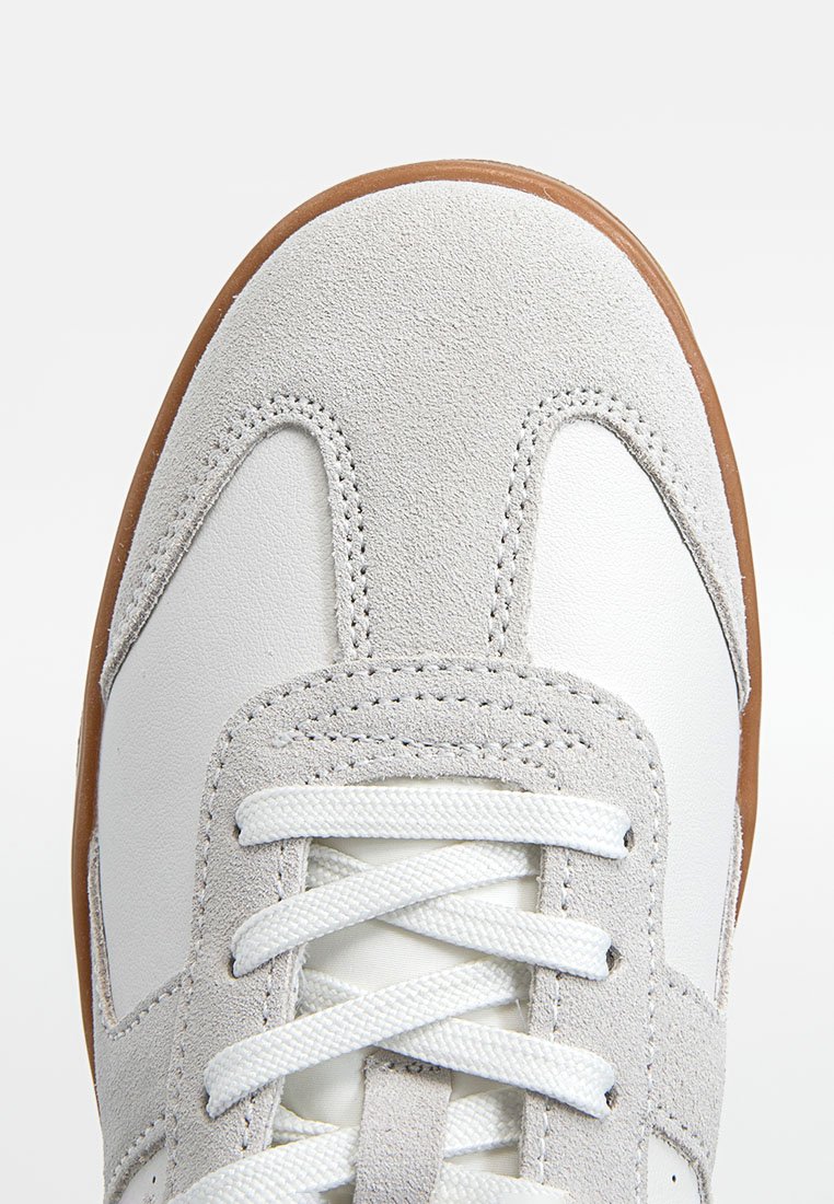 Szare zamszowe i białe skórzane sneakersy z okrągłym noskiem, płaskimi białymi sznurowadłami oraz jasnobrązową gumową podeszwą. Dodatkowe przeszycia na górze.