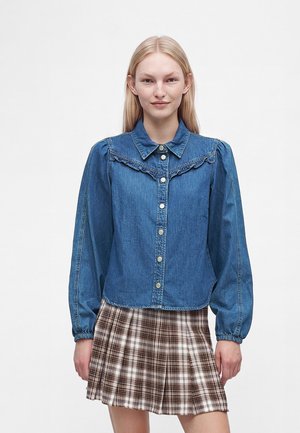 Ung kvinde iført en blå denim skjorte med knapper og flæsedetalje samt et brunt ternet plisseret skørt, stående mod en hvid baggrund.
