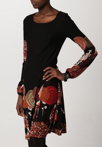 Femme portant une robe noire à manches longues avec des motifs abstraits colorés circulaires et géométriques sur les manches et l'ourlet.