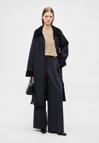 Hosszú sötétkék kabát fekete gallérral, bézs felsővel és széles szárú sötétkék nadrággal. Az öltözékhez kis fekete kézitáska és hegyes orrú cipők tartoznak.