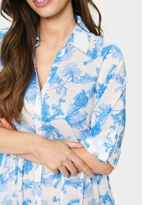 Vit blus med blå blommönster. Har krage, knappstängning fram och uppvikta ärmar. Tyget är lätt och semitransparent.